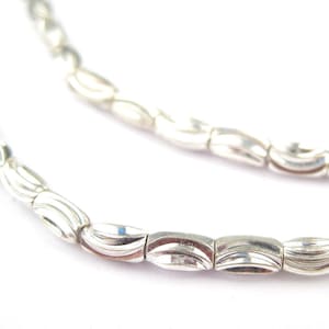 May include: A silver chain necklace with a wavy, textured design. The chain is made of small, linked oval-shaped beads.