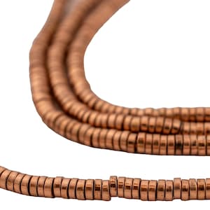 以下が含まれることがあります： 複数のストランドを持つ銅製のネックレスのクローズアップ。ネックレスは、小さくて丸い銅製のビーズでできています。ビーズは光沢のある金属仕上げで、白い背景に対して流れるような曲線模様に配置されています。