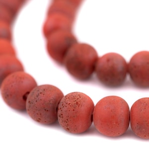 May include: Close-up of a strand of red-orange beads. The beads are round, with a matte finish and a slightly speckled texture. The beads are strung together, creating a curved line against a white background.