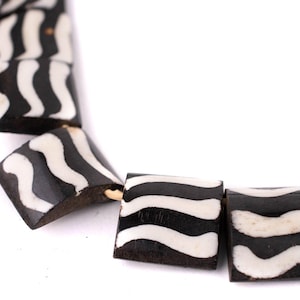 May include: Close-up of a necklace featuring square beads with a black and white zebra stripe pattern. The beads are strung together, showcasing a unique design. The necklace is set against a white background.