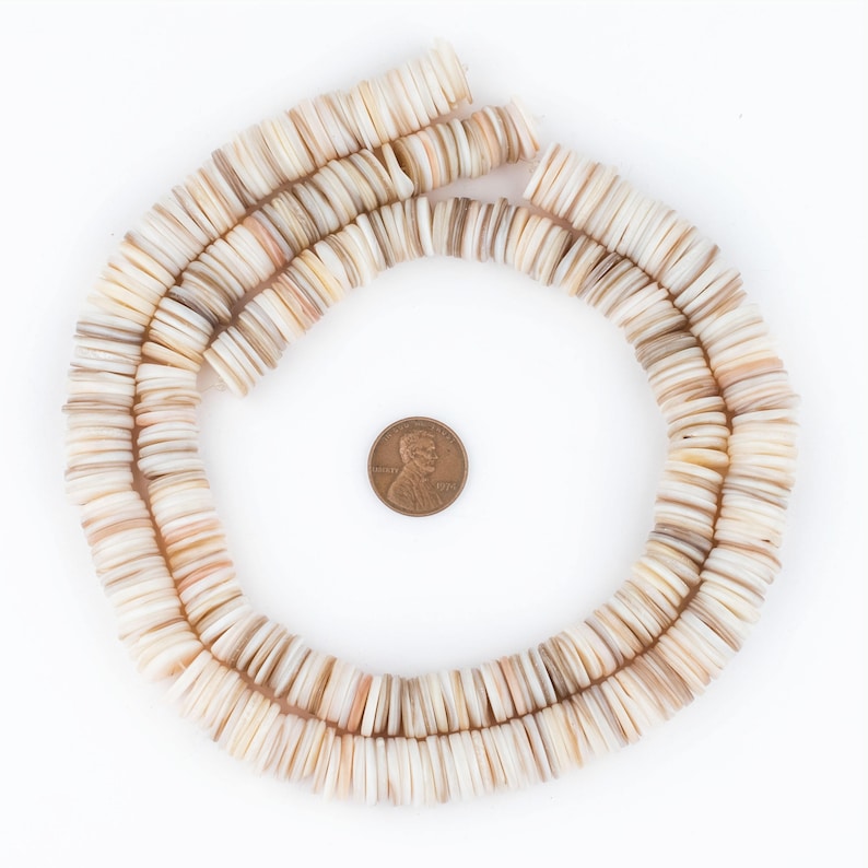 May include: A necklace of flat, round, off-white and tan shell beads arranged in a circular pattern. A U.S. penny is in the center for size comparison. The beads have a natural, organic appearance, with variations in color and texture.