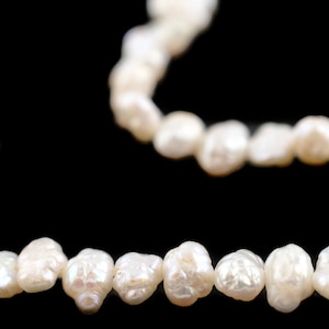 Puede incluir: Primer plano de un collar de perlas blancas de forma irregular sobre un fondo negro. Las perlas son luminosas y tienen un aspecto natural y orgánico. El collar es probablemente para joyería.