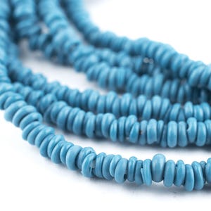 Könnte beinhalten: Nahaufnahme von mehreren Strängen türkisfarbener Perlen. Die Perlen sind klein, unregelmäßig geformt und aufgefädelt. Der Hintergrund ist weiß und hebt den leuchtenden Blauton der Perlen hervor. Die Perlen scheinen aus einem natürlichen Material zu bestehen.