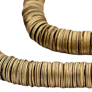 May include: Close-up of a necklace made of numerous small, round, flat, golden-colored beads. The beads are strung together, forming a circular shape. The necklace has a natural, earthy aesthetic, with a warm, neutral color palette.
