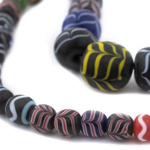 Könnte beinhalten: Eine Kette aus dunklen Perlen mit Streifenmustern in Weiß, Blau, Rot und Gelb. Die Perlen sind schwarz und rund, mit matter Oberfläche. Die Perlen sind zu einer Halskette oder einem Armband aufgereiht. Die Perlen haben einen Durchmesser von etwa 1 cm.