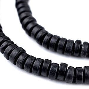 Puede incluir: Primer plano de un collar de cuentas negras. Las cuentas son cilíndricas con una textura ligeramente rugosa y están ensartadas para formar un collar o pulsera. El fondo es blanco, resaltando las cuentas oscuras.
