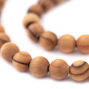 Pode incluir: Close-up de um fio de contas de madeira. As contas são redondas, castanho-claras e têm padrões de grão castanho-escuro. As contas são enfiadas, formando uma linha curva contra um fundo branco. As contas de madeira natural são provavelmente usadas para fazer joias.