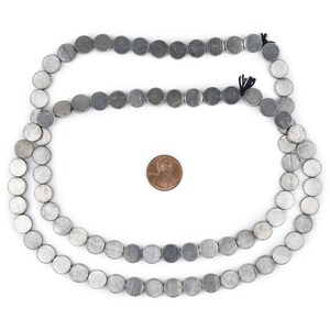 Pu&ograve; includere: Una collana di perline rotonde argentate, di circa 10 mm di diametro, disposte in cerchio su uno sfondo bianco. Un centesimo &egrave; posto al centro del cerchio per il confronto delle dimensioni.