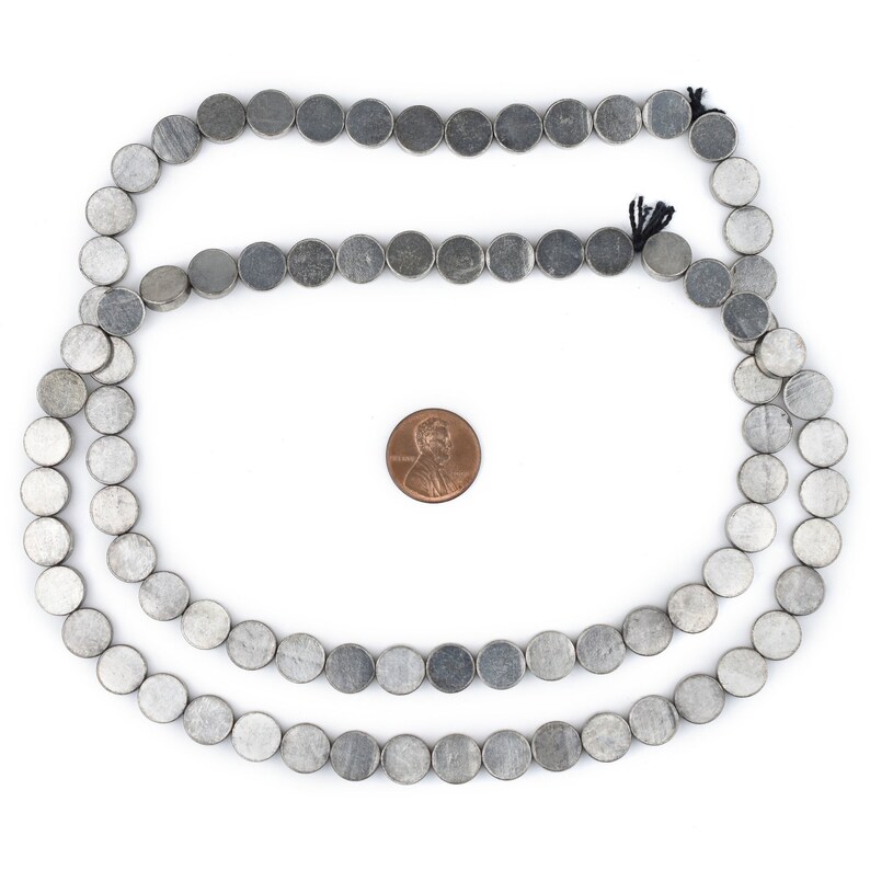Pu&ograve; includere: Una collana di perline rotonde argentate, di circa 10 mm di diametro, disposte in cerchio su uno sfondo bianco. Un centesimo &egrave; posto al centro del cerchio per il confronto delle dimensioni.