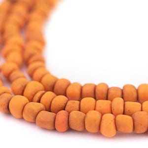 May include: Close-up of three strands of orange beads. The beads are a muted orange color and have a slightly irregular, textured surface. The beads are strung together, creating a necklace or bracelet. The background is white.