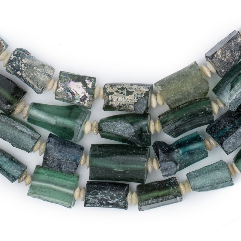 May include: A close-up of a necklace made of green and blue glass beads. The beads are irregular in shape and have a mottled appearance. The necklace is strung on a white cord.