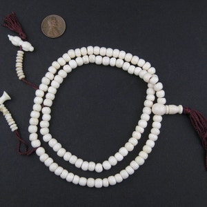 May include: Ivory-colored prayer beads with a dark red tassel and decorative end pieces. The beads are arranged in two strands, with a penny for size comparison. The background is a solid black.