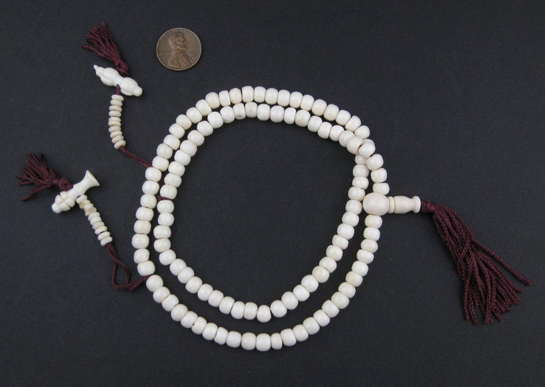 May include: Ivory-colored prayer beads with a dark red tassel and decorative end pieces. The beads are arranged in two strands, with a penny for size comparison. The background is a solid black.