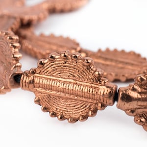 Op de afbeelding: Een close-up van een koperkleurige ketting met ingewikkelde, gedetailleerde, ovale kralen. De kralen hebben een getextureerde, gehamerde afwerking.