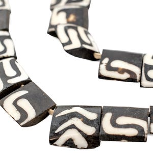 Puede incluir: Un collar hecho de cuentas de hueso negro y blanco con un patrón geométrico. Las cuentas son de forma cuadrada y tienen un acabado liso y pulido.
