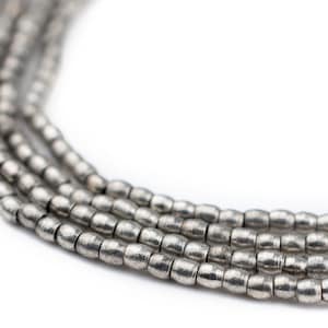 May include: A close-up of a silver metal bead necklace. The beads are small and round, and they are strung together in a simple pattern. The necklace is lying on a white surface.