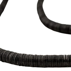 Op de afbeelding: Close-up van een zwarte, gebogen ketting. De ketting is gemaakt van kleine, ronde, donkergrijze kralen, waardoor een textuur ontstaat. Het ontwerp is eenvoudig en elegant, geschikt voor diverse stijlen.