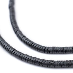 Puede incluir: Primer plano de dos hilos de cuentas planas en forma de disco, de color gris oscuro. Las cuentas están ensartadas, formando un collar o una pulsera. El fondo es blanco liso, resaltando la textura y el color de las cuentas.