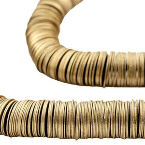 May include: A gold-colored necklace composed of numerous small, circular, flat metal pieces strung together. The necklace is arranged in a curved shape against a white background, showcasing its design and texture.