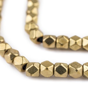 以下が含まれることがあります： 面取りされた幾何学的な形をした金色の金属ビーズのストランド。ビーズは黒いコードに糸で通されています。