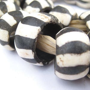 May include: A close-up of a string of black and white striped beads. The beads are made of bone or ivory and are strung on a natural fiber cord.