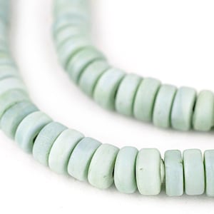 May include: Close-up of a necklace made of light green, flat, round beads. The beads are strung together, creating a curved line against a white background. The necklace appears to be handmade.