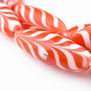 May include: Close-up of red and white striped glass beads. The beads are oval shaped and have a swirl pattern.