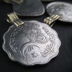 May include: Silver pendant with intricate Arabic calligraphy and a crescent moon and star design. The pendant has a gold-colored loop for attaching a chain.