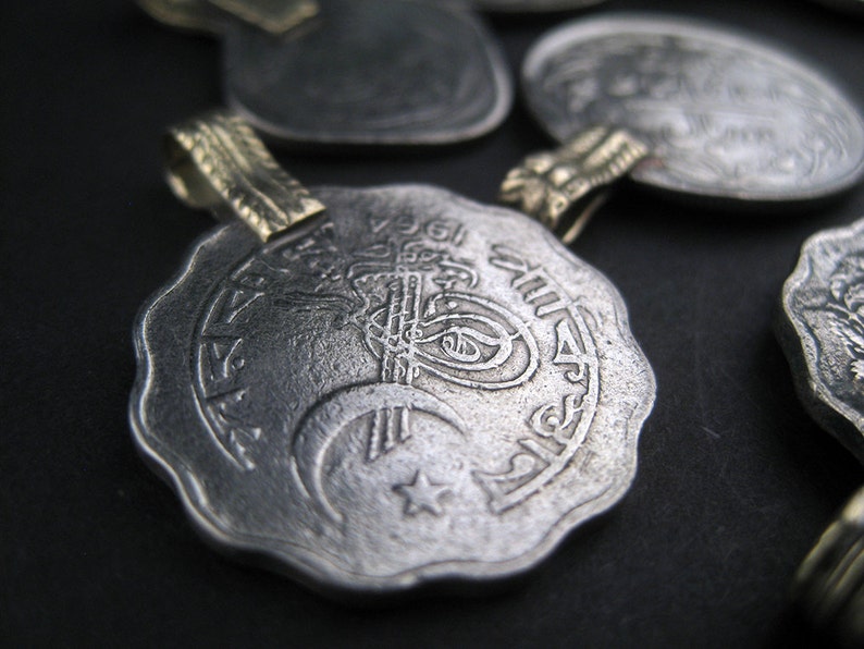May include: Silver pendant with intricate Arabic calligraphy and a crescent moon and star design. The pendant has a gold-colored loop for attaching a chain.
