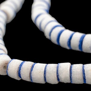 Pode incluir: Close-up de um colar de contas brancas com listras azuis. As contas cilíndricas são enfiadas, criando uma linha curva sobre um fundo preto. As contas parecem ser feitas de um material poroso.