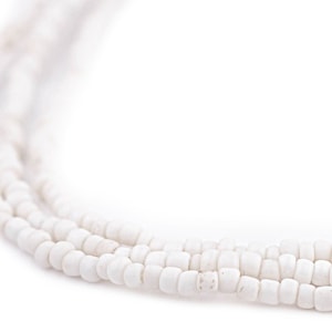 Könnte beinhalten: Nahaufnahme einer weißen Perlenkette. Die Halskette besteht aus mehreren Strängen kleiner, rechteckiger Perlen. Die Perlen sind einfarbig, undurchsichtig weiß. Die Halskette wird vor einem weißen Hintergrund gezeigt.
