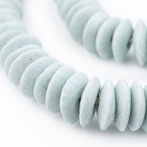 May include: Close-up of a necklace with light blue, disc-shaped beads. The beads are strung together, forming a curved line against a white backdrop. The necklace appears to be made of a textured material.