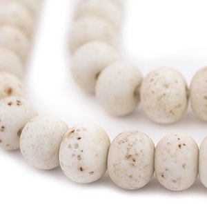 May include: Close-up of a strand of off-white, oval-shaped beads. The beads have a speckled, aged appearance with small brown spots. The beads are strung together, suggesting they are for jewelry making or crafting.