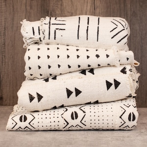 May include: A stack of four cream-colored, patterned textiles. Each textile features a different black geometric design, including lines, triangles, and dots. The textiles are stacked neatly, with visible fringe on the edges. The background is a wooden surface.