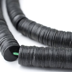 May include: A close-up shot of a black necklace composed of flat, circular beads. The beads are strung together, forming a flexible strand. The necklace is set against a white backdrop, accentuating the dark colour and texture of the beads.