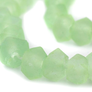 May include: A string of pale green, faceted beads. The beads have a slightly frosted appearance and a geometric shape. The beads are arranged in a curved line against a white background. The beads are likely for jewellery making.