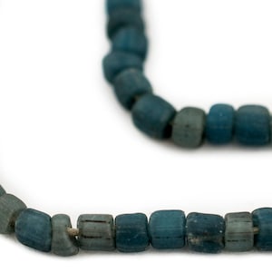 Puede incluir: Primer plano de un collar con cuentas azul-verdosas en forma de barril. Las cuentas tienen un aspecto ligeramente desgastado, con variaciones de color de azul-verdoso claro a oscuro. El collar se muestra sobre un fondo blanco.