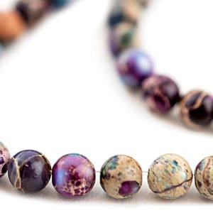 May include: A strand of gemstone beads in shades of purple, blue, and tan. The beads are round and have a marbled appearance. The beads are strung together, creating a necklace or bracelet.