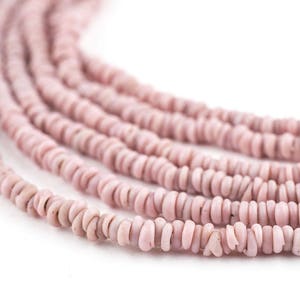 May include: Close-up of several strands of pink shell beads. The beads are small, flat, and irregularly shaped, with a slightly rough texture. The color is a soft, muted pink, and the beads are strung together, creating a flowing, organic look.