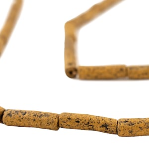 May include: A necklace featuring cylindrical beads with a speckled, textured surface. The beads are a warm, earthy yellow-brown color with darker brown speckles. The necklace is displayed against a white background.