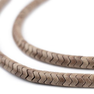 May include: Close-up of a light brown, textured necklace with a repeating zigzag pattern. The necklace is made of a flexible material and appears to be worn as jewelry. The background is white.