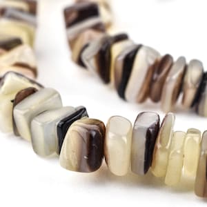 May include: A close-up of a necklace made of irregularly shaped, polished beads. The beads are a mix of neutral colors, including cream, brown, and black. The necklace is displayed against a white background.