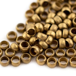 May include: Close-up of a pile of small, cylindrical brass beads. The beads have a rounded shape with a hole running through the center. The beads are scattered on a white surface, with some in focus and others blurred.