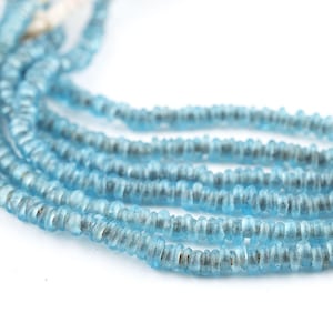 Puede incluir: Primer plano de varios hilos de cuentas de piedras preciosas azul claro. Las cuentas son pequeñas, redondas y tienen una forma ligeramente irregular. El fondo es blanco, resaltando el color y la textura de las cuentas.