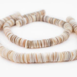 May include: A necklace made of flat, round, shell beads in shades of cream, tan, and brown. The beads are strung together, forming a curved shape against a white background. The necklace appears to be handmade and natural.