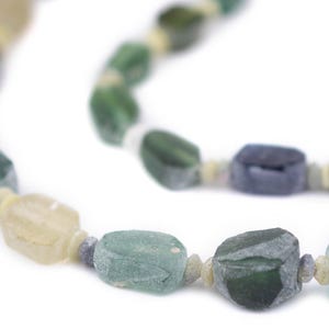 May include: A close-up of a necklace with multi-colored, rectangular beads. The beads are in shades of green, blue, and yellow, strung together with small, light-colored spacers. The necklace is set against a white background.
