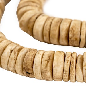 May include: Close-up of a necklace made of light beige, flat, circular beads. The beads have a natural, organic look with variations in color and texture. The necklace is displayed against a white background.
