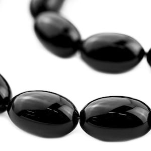 Könnte beinhalten: Nahaufnahme einer Halskette mit großen, glänzenden schwarzen Perlen. Die oval geformten Perlen sind aufgereiht und bilden eine geschwungene Linie vor einem weißen Hintergrund. Die Perlen reflektieren das Licht und wirken poliert.