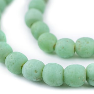 以下が含まれることがあります： マット仕上げの薄緑色のビーズ2連のクローズアップ。ビーズは丸みを帯びた形で、わずかに平らな形とテクスチャ加工された表面を持っています。ビーズは一緒に紐で繋がれており、ネックレスまたはブレスレットを作成しています。