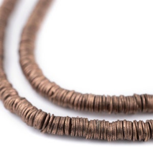 Peut inclure: Gros plan sur deux rangs d'un collier composé de petites perles rondes de couleur cuivrée. Les perles sont enfilées ensemble, créant un effet texturé et superposé. Le fond est blanc uni, mettant en valeur les détails du collier.